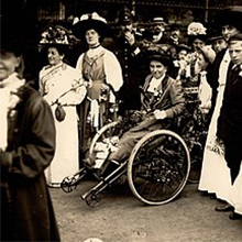 Rosa May Billinghurst, dressed in long dress and hat, sits in her adapted tricycle. She is surronded by a group of women Suffragettes, looking towards the camera. 